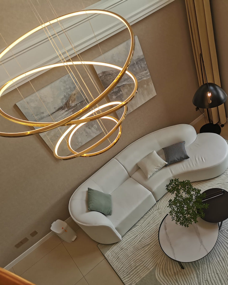 Top-down view of illuminated gold rings above a curved sofa, showing how the chandelier becomes art from every floor and viewing angle.