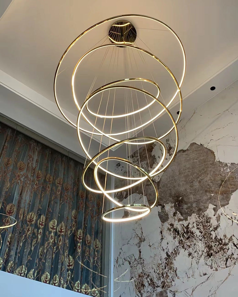 Fully illuminated entryway chandelier with overlapping gold rings, creating a rich cascade of warm light against marble and patterned drapery.