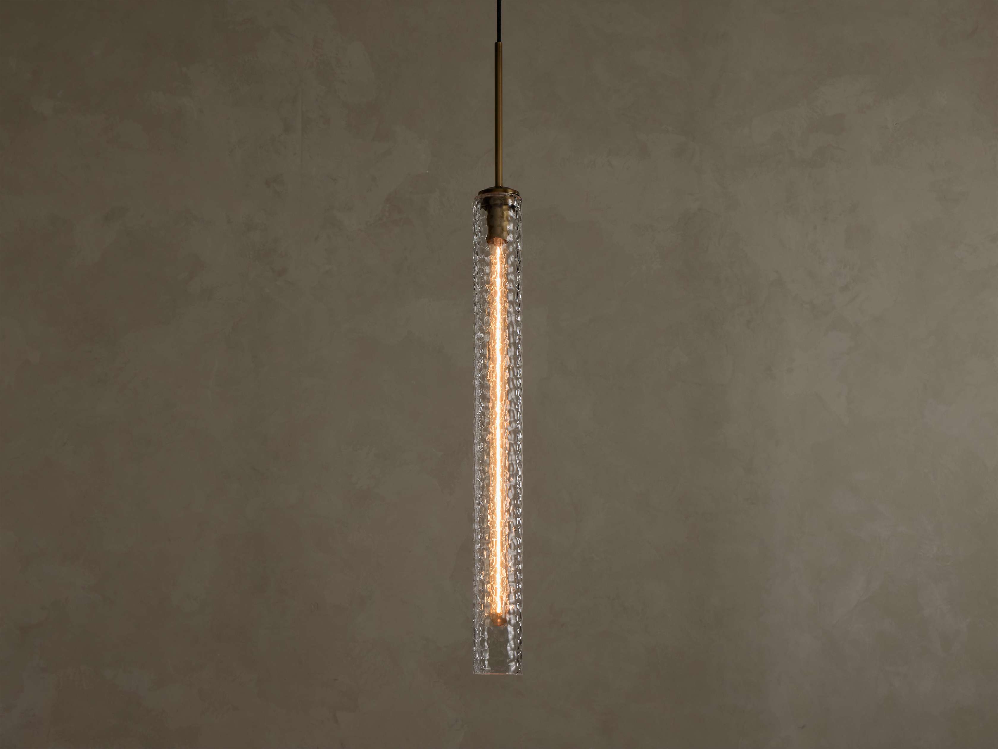 Vertical view of a lit pendant light with a clear textured glass cylinder, a warm filament bulb, and a brass-colored top fixture.