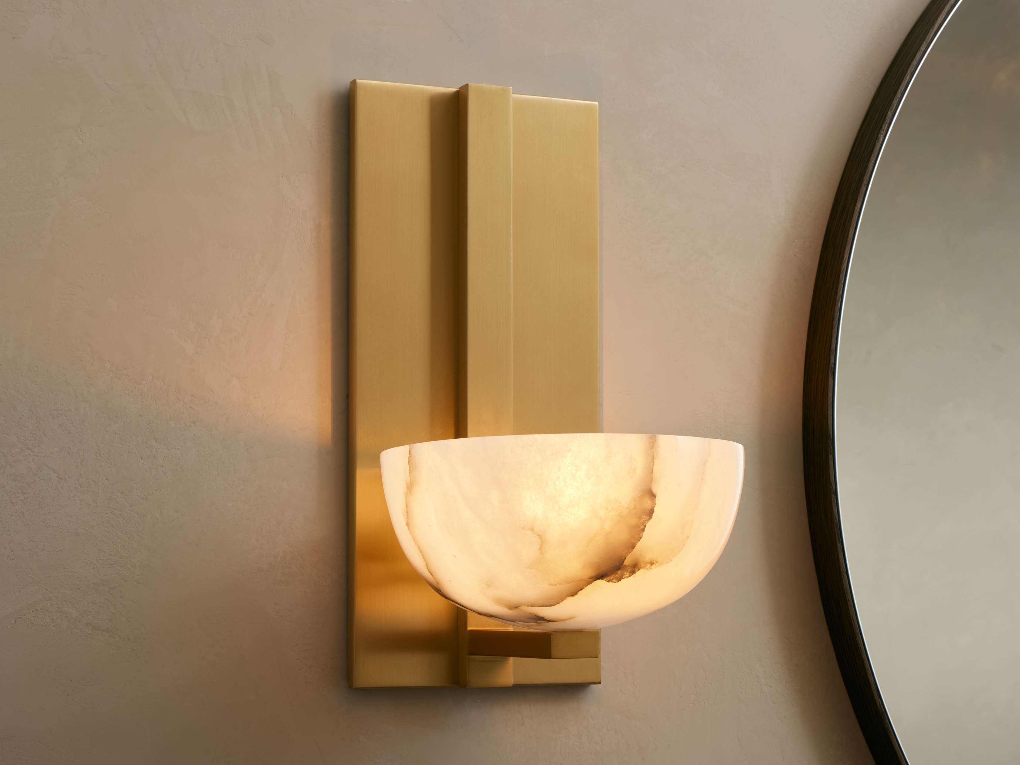Belmira Sconce: Brass-finished wall light with glowing, bowl-shaped alabaster shade on a textured wall next to a mirror.