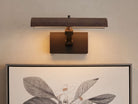 Abel Picture Light Sconce, a dark bronze curved light bar with warm LED light, mounted on a wall above a framed botanical artwork.