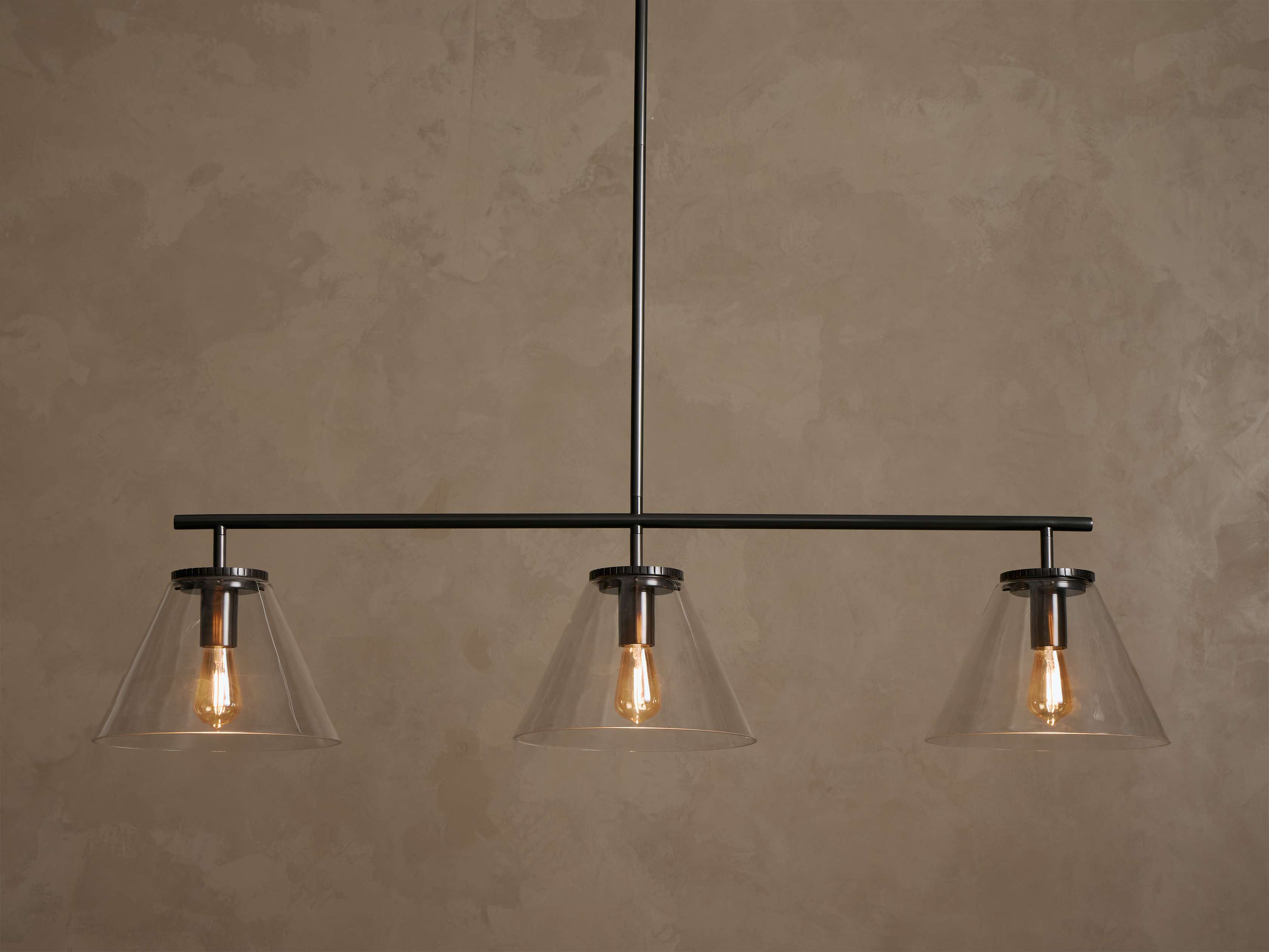 Front view of a black linear pendant light fixture with three clear conical glass shades and exposed warm Edison bulbs.