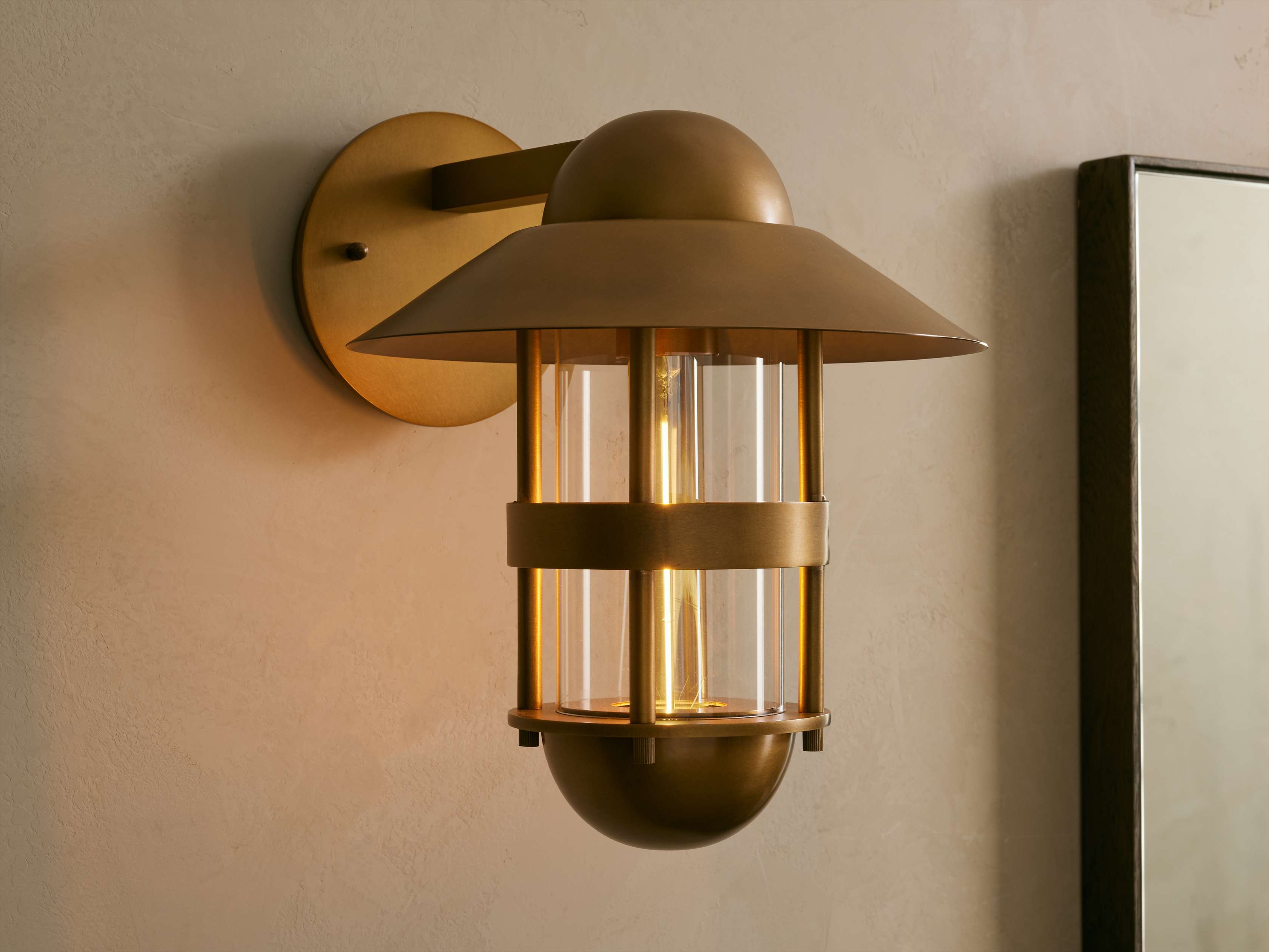 Illuminated Hyannis Outdoor Sconce, antique brass, nautical style with glass housing and visible bulb, on a light wall.